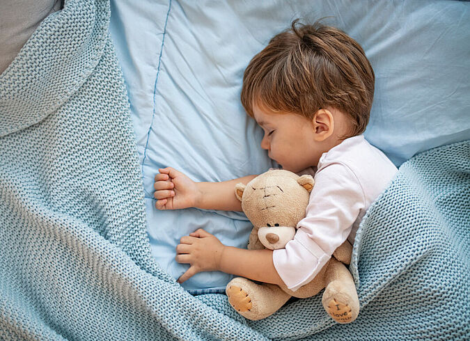 Ein Kleinkind schläft mit Kuschelbär im Arm in einem Bett.